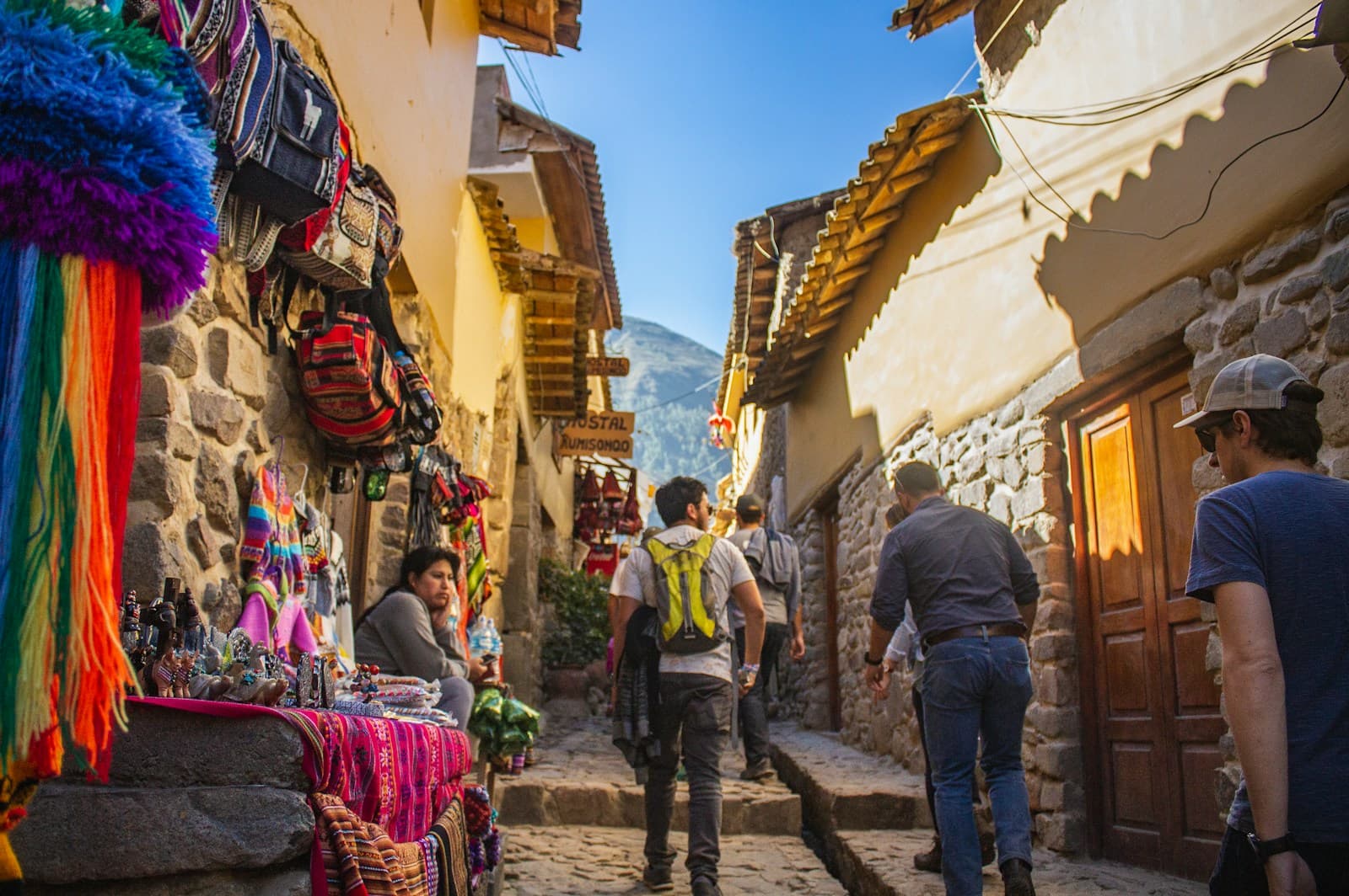 Travelers with backpacks walking up a stone street in Ollantaytambo, past stalls of colorful Andean textiles.